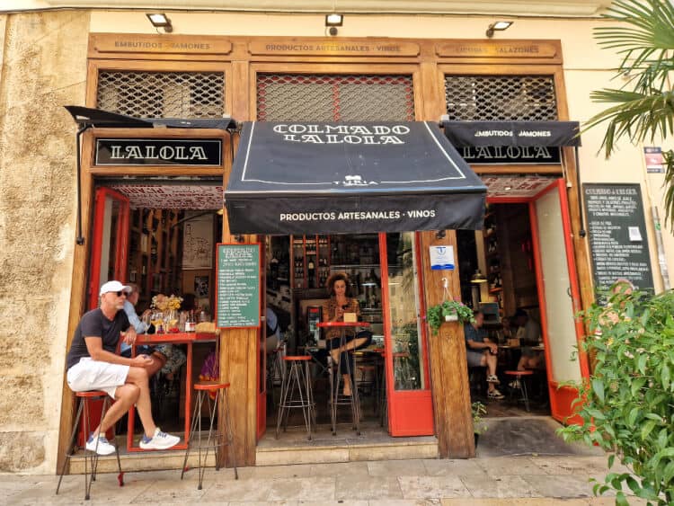 La entrada del Colmado Lalola, un establecimiento acogedor en Valencia, con clientes disfrutando de tapas al aire libre. Un lugar que refleja el encanto de la vida urbana y la gastronomía local valenciana.