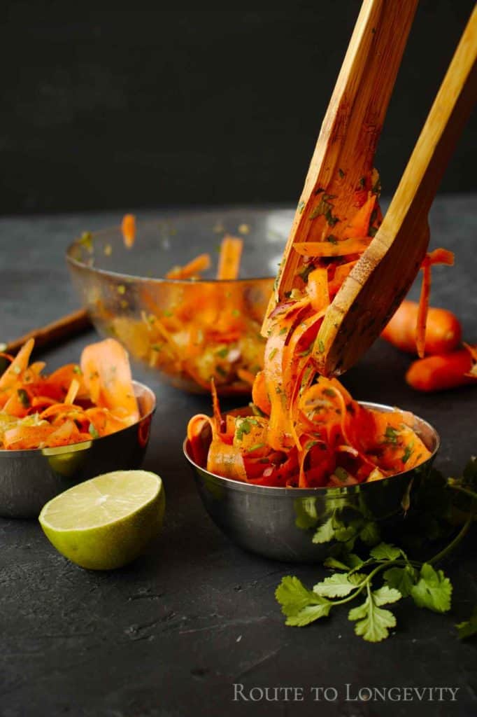 A bowl of Asian carrot salad with julienned carrots, cilantro, and dressing being served with wooden tongs. A lime half and additional salad in a metal bowl are visible in the foreground.