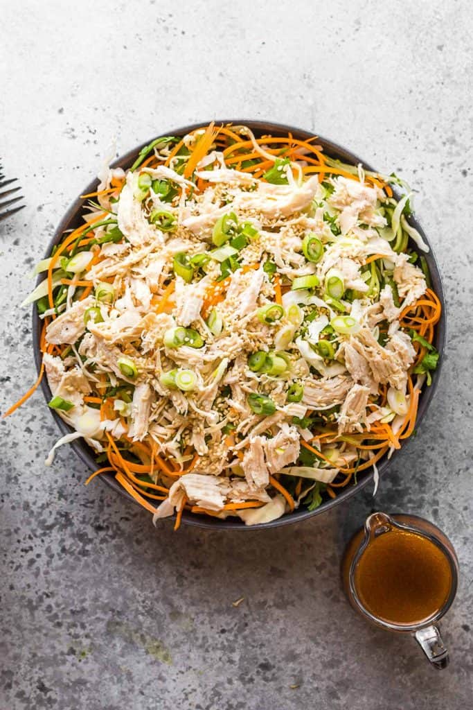 A bowl of Asian chicken salad with shredded chicken, julienned carrots, green onions, and sesame seeds, served with a small pitcher of dressing on the side.