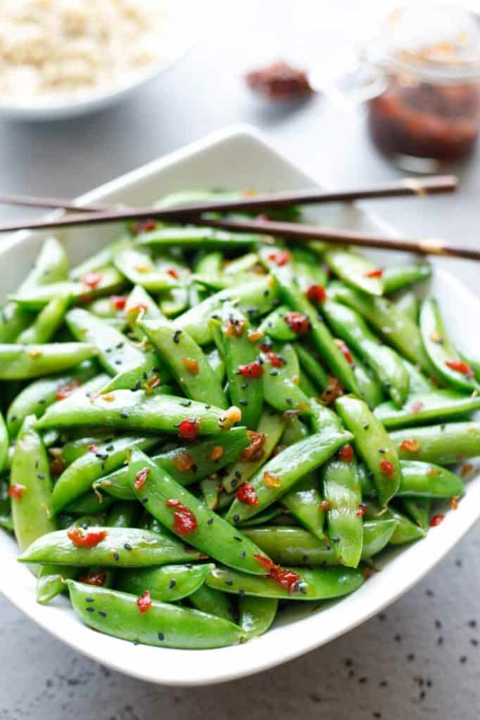 A bowl of Asian-style sugar snap peas stir-fried with red chili flakes and sesame seeds, served with chopsticks. A small jar of sauce and a bowl of rice are blurred in the background.