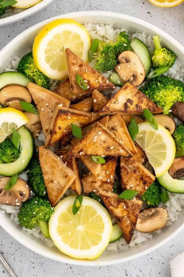 A bowl of Asian marinated tofu served with broccoli, mushrooms, cucumber, and lemon slices on a bed of rice. Garnished with green onions and sesame seeds.