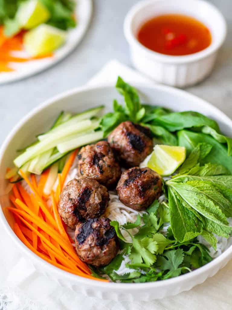 A bowl of Vietnamese bún chả with grilled pork meatballs, vermicelli noodles, sliced cucumbers, carrots, fresh herbs, and a lime wedge. A small bowl of dipping sauce is in the background.