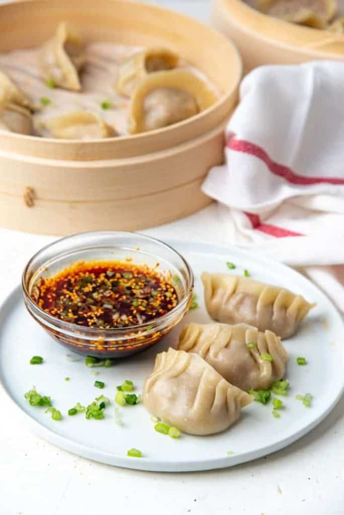 A plate with three chicken dumplings garnished with green onions, served with a bowl of dipping sauce. A bamboo steamer filled with additional dumplings is in the background.