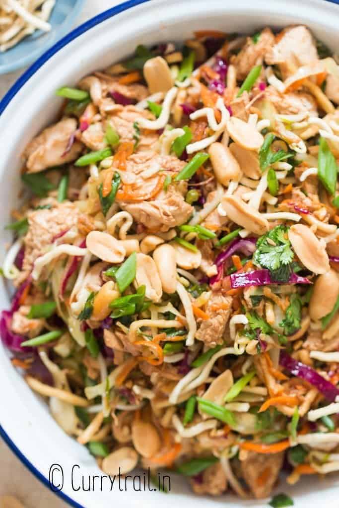 A vibrant Chinese chicken salad featuring shredded chicken, crunchy peanuts, and fresh vegetables like red cabbage, carrots, and green onions, all mixed with crispy noodles. The dish is garnished with cilantro and presented in a white bowl with a blue rim.