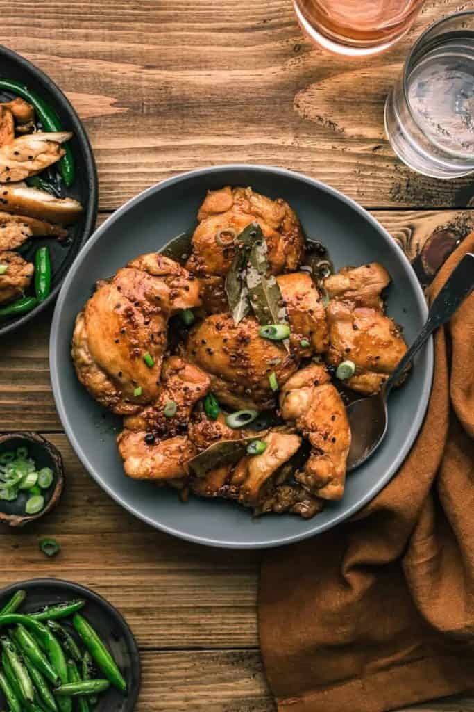 A plate of Filipino chicken adobo, featuring tender chicken pieces braised in a savory soy sauce and vinegar marinade, garnished with bay leaves and green onions. The dish is served on a gray plate with a side of sautéed green beans, and set on a rustic wooden table alongside a glass of water and a brown napkin.