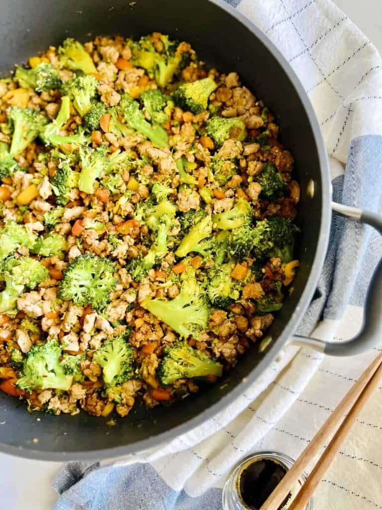 A pan filled with ground pork stir-fry with broccoli, carrots, and cashews. The pan is placed on a cloth, with chopsticks and a small jar of sauce nearby.