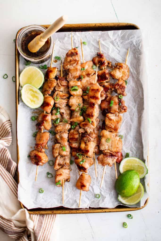 A tray of Japanese yakitori skewers, garnished with green onions and served with lime wedges. A small jar of sauce with a basting brush is placed on the tray.