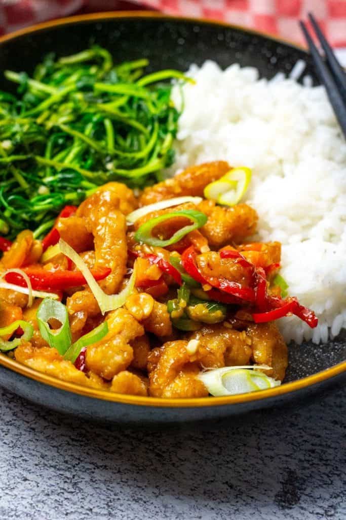 A bowl of Kung Pao chicken served with steamed white rice and sautéed greens. The dish includes crispy chicken pieces, sliced red and green peppers, and is garnished with green onions.