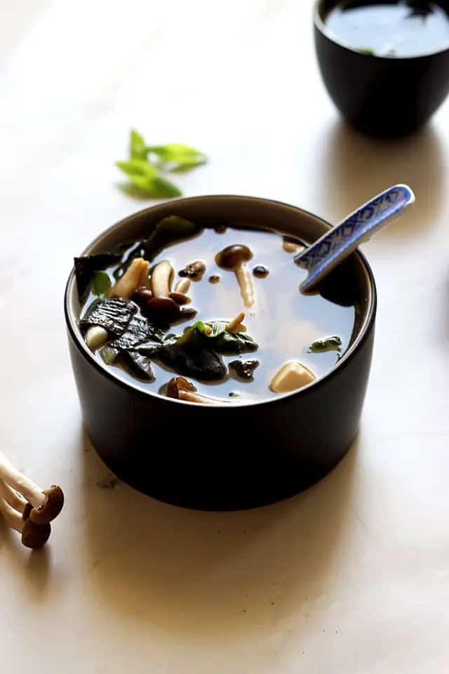 A bowl of miso soup containing seaweed, tofu, mushrooms, and green onions, with a ceramic spoon resting inside. Another smaller bowl is visible in the background.