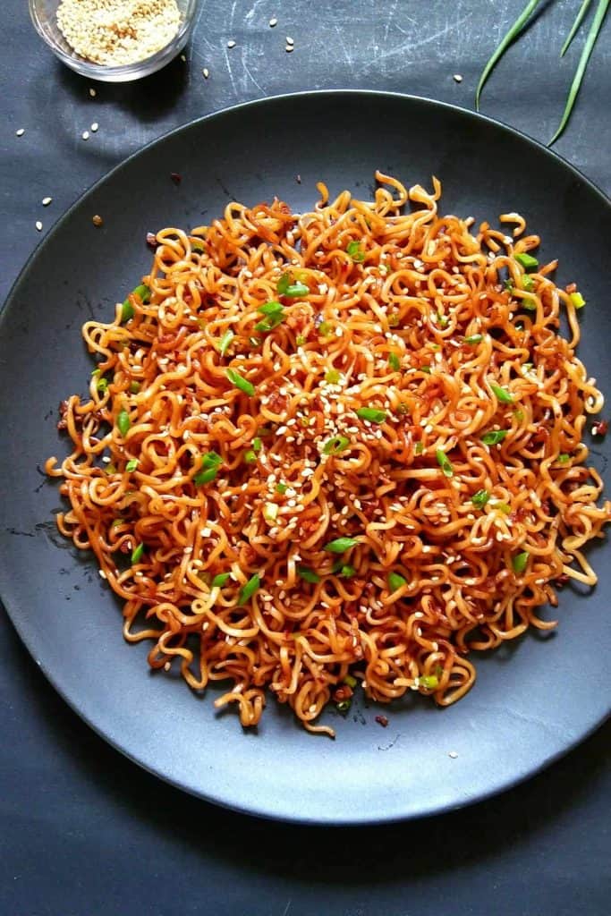 A black plate filled with sesame garlic ramen noodles, garnished with green onions and sesame seeds. A small bowl of sesame seeds is visible in the background.