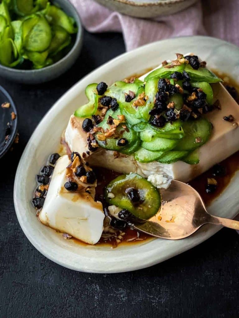 A plate of silken tofu topped with pickled cucumber slices, black beans, and a savory sauce. A spoon is lifting a piece of the tofu, and a bowl of additional pickled cucumbers is visible in the background.