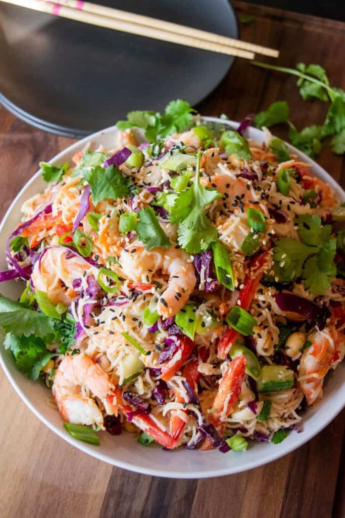 A bowl of Thai shrimp noodle salad, featuring vermicelli noodles, shrimp, red cabbage, bell peppers, green onions, cilantro, and sesame seeds. Chopsticks and a plate are visible in the background.
