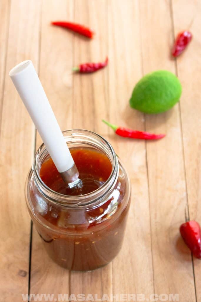 A jar of Thai sweet chili lime sauce with a white spoon inside, placed on a wooden surface. Surrounding the jar are red chili peppers and a lime, highlighting the key ingredients.