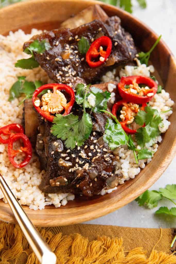 A bowl of Asian beef short ribs served on a bed of fluffy rice. The tender ribs are garnished with sesame seeds, fresh cilantro, and sliced red chili peppers, adding a pop of color and heat. A gold-colored fork is placed next to the bowl, which sits on a textured fabric napkin.