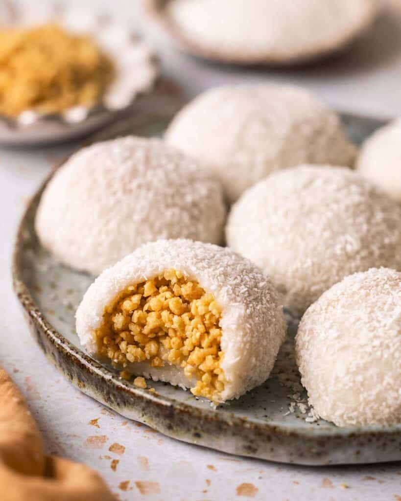 A plate of coconut peanut mochi, with one mochi partially bitten to reveal the peanut filling. Additional mochi and a small dish of crushed peanuts are visible in the background.