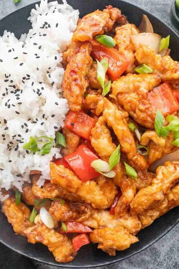 A plate of dragon chicken served with steamed white rice sprinkled with black sesame seeds. The chicken is crispy, coated in a sweet and spicy sauce, and garnished with chopped green onions and red bell peppers.