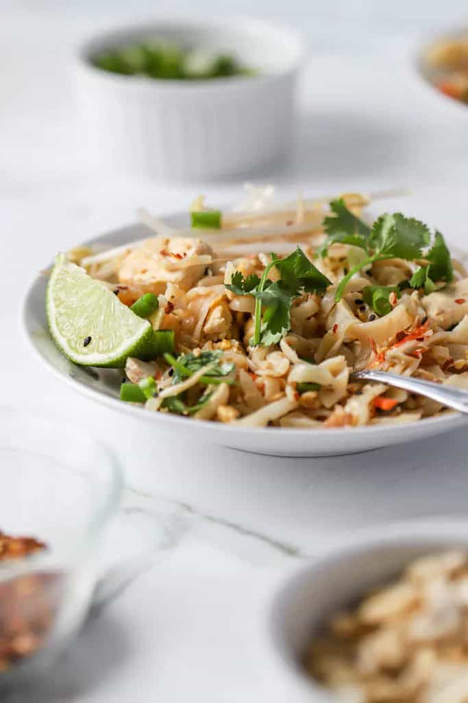 A bowl of flat rice noodles topped with a creamy peanut sauce, garnished with fresh cilantro, green onions, and bean sprouts. A lime wedge is placed on the side for added zest.