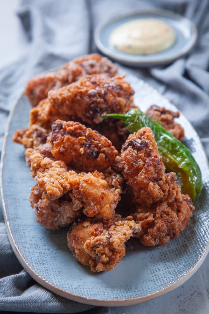 A plate of Japanese karaage, featuring crispy fried chicken pieces garnished with a green pepper. A small dish of dipping sauce is blurred in the background.