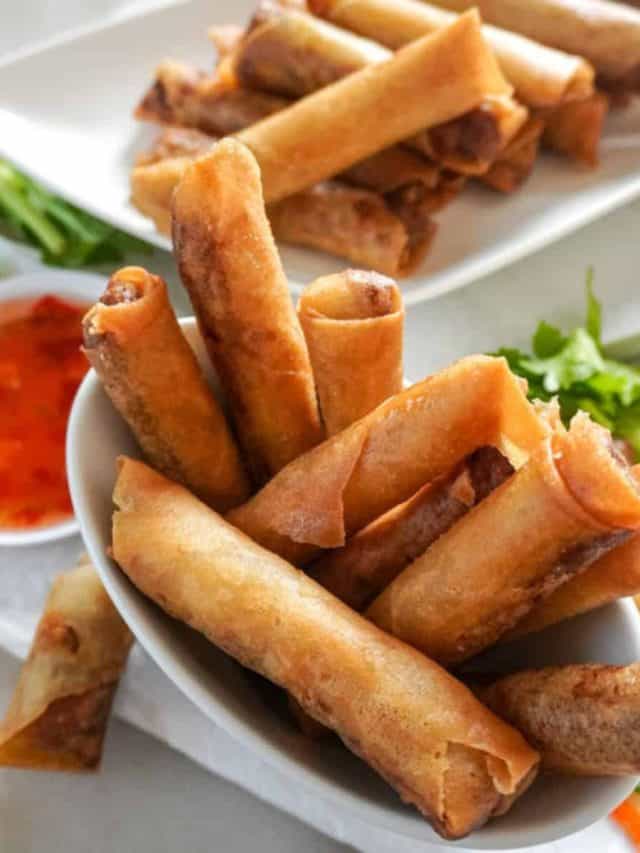 A bowl and a plate filled with crispy Filipino lumpia, accompanied by a small bowl of sweet chili dipping sauce, garnished with fresh herbs.