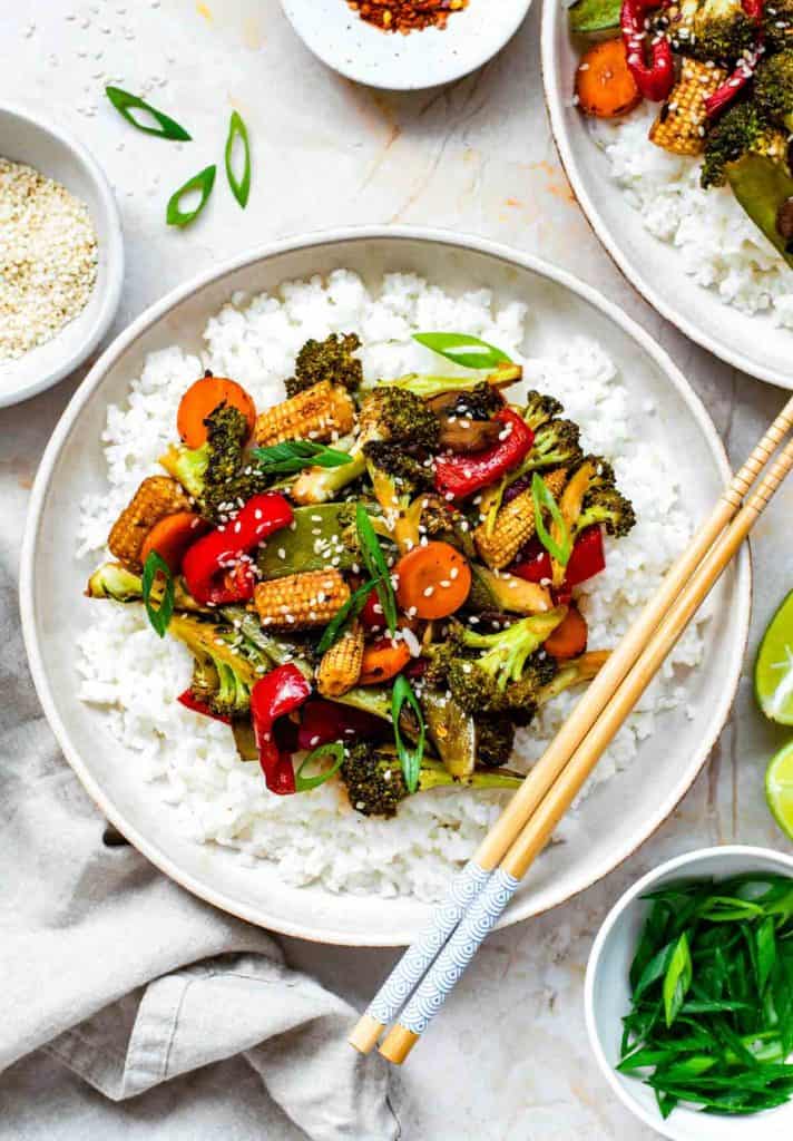 A bowl of Pad Pak, a Thai vegetable stir-fry, served over white rice with broccoli, baby corn, bell peppers, carrots, and garnished with sesame seeds and green onions. Chopsticks rest on the side of the bowl.