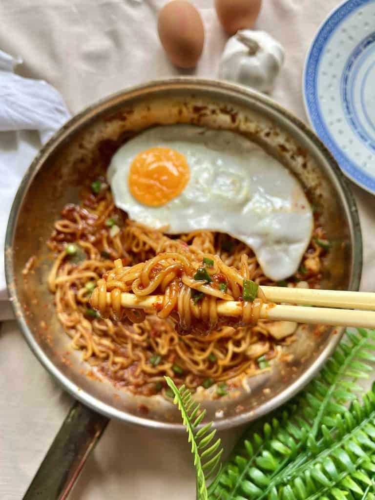 A pan of spicy gochujang noodles garnished with green onions and topped with a fried egg with a perfectly cooked yolk. In the foreground, chopsticks are lifting a bite of the saucy noodles.