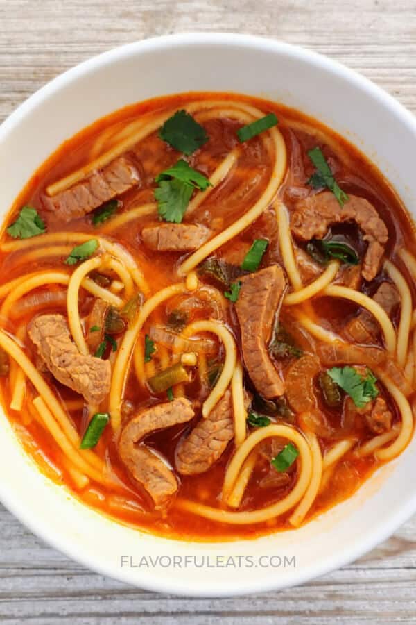 A bowl of spicy Thai beef noodle soup with tender beef slices, noodles, and fresh herbs in a rich red broth.