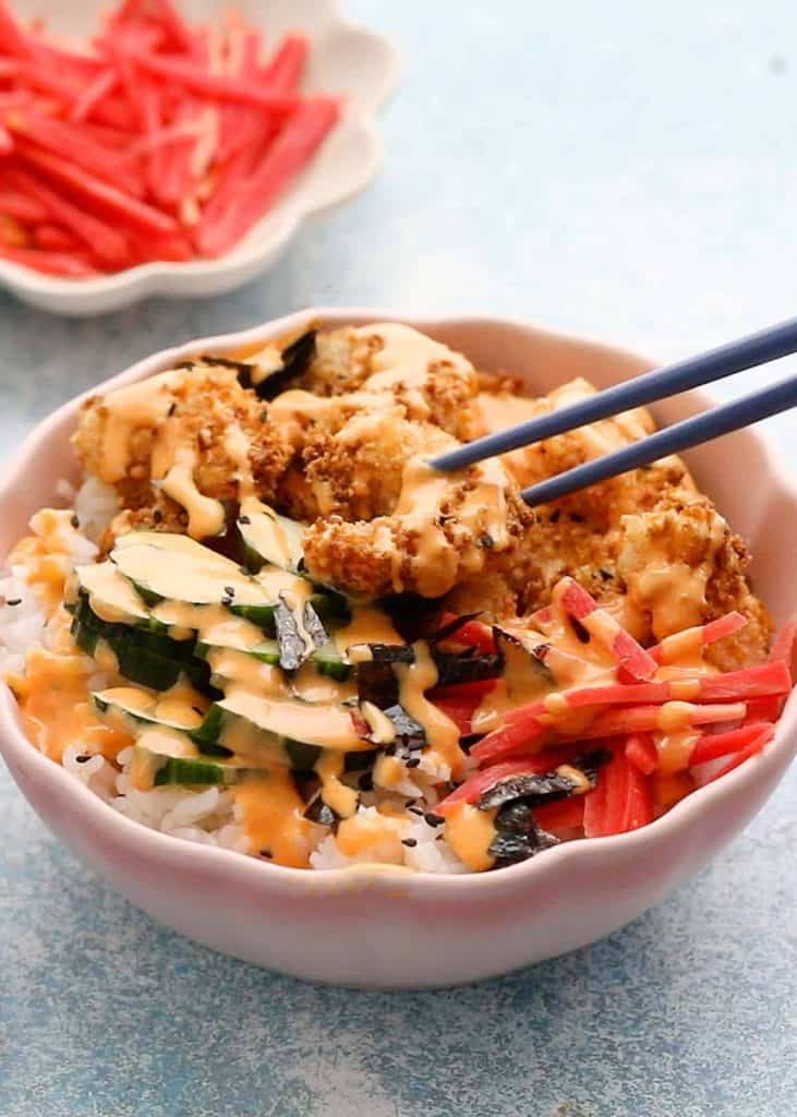 A sushi bowl with rice, crispy fried pieces, sliced cucumber, pickled ginger, and seaweed, drizzled with a creamy sauce. Chopsticks are picking up a piece from the bowl, with extra pickled ginger in the background.