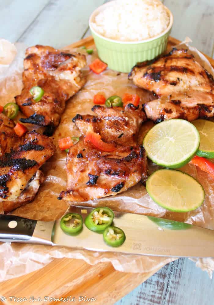 Grilled Thai chicken thighs with a caramelized, charred exterior, served with lime wedges, sliced green chilies, and red bell peppers. A green ramekin filled with steamed white rice is placed in the background, and a knife rests on the parchment paper-lined wooden board for easy serving.