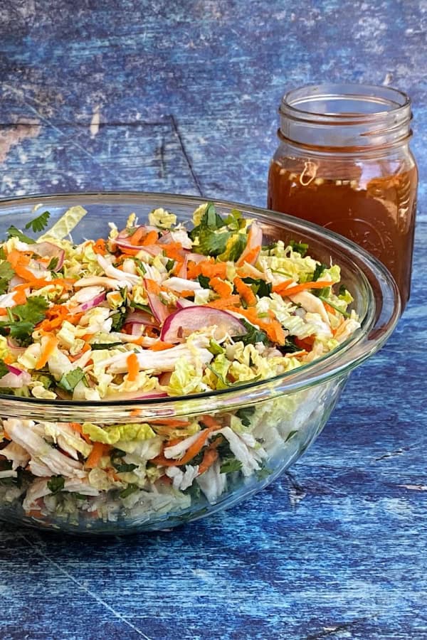 A glass bowl filled with Vietnamese chicken salad, featuring shredded chicken, napa cabbage, carrots, red onions, and fresh herbs like cilantro and mint. A jar of dressing is placed in the background, and the salad is set against a textured blue background.